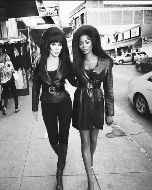 Black and white photo of Cher and Naomi Campbell walking arm in arm on a city street in matching jackets and dramatic hairstyles.