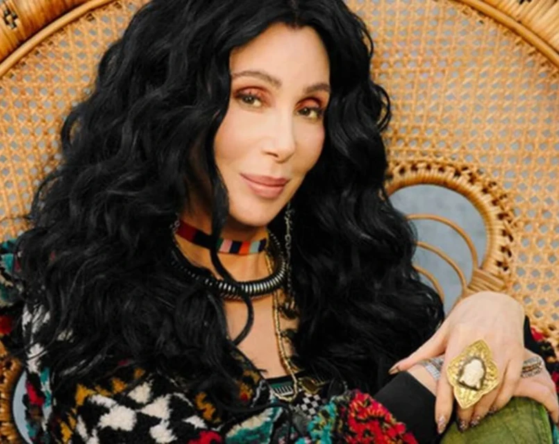 Portrait of an adult woman with long, dark wavy hair, wearing multiple necklaces and a large gold ring, seated in front of a woven chair.