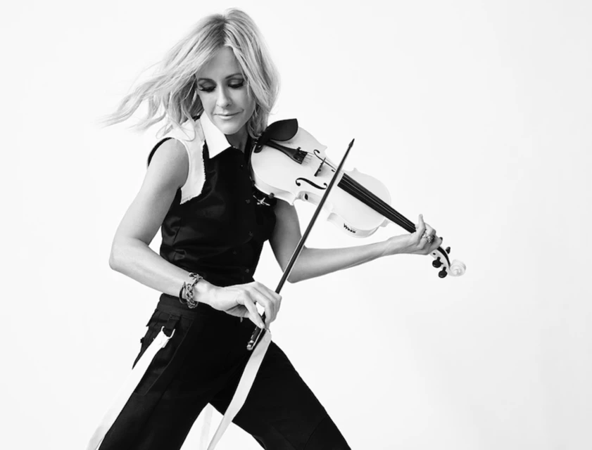 A woman with fair hair is playing a violin in a black and white portrait, wearing a black sleeveless top and black pants with white stripes.