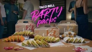 A table laden with Taco Bell party packs, surrounded by a blurred group of people, hints at a casual gathering and fast-food catering.