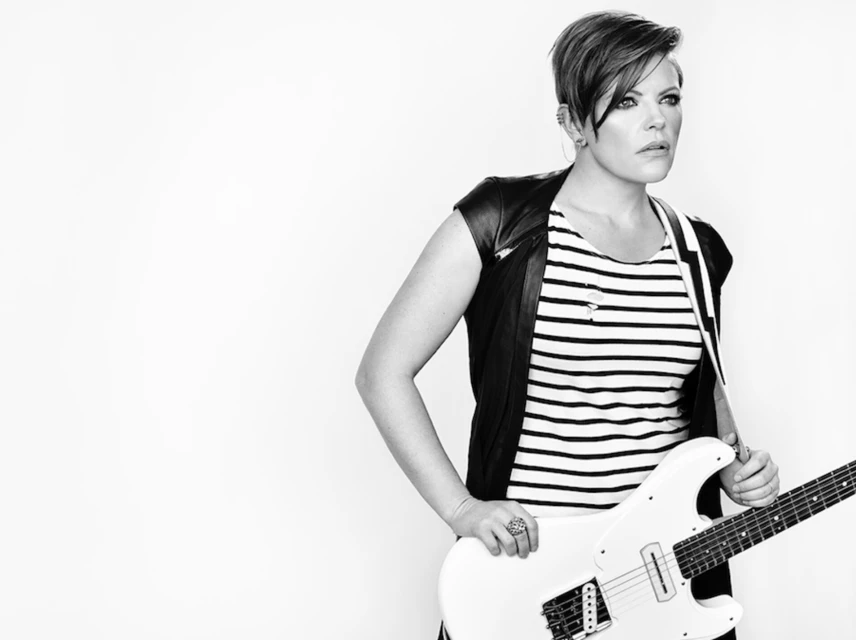 Monochrome portrait of a woman with a short hairstyle in a striped top, holding a white electric guitar against a plain white backdrop.