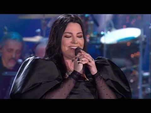 Female singer in concert, wearing a black dress, performs on stage with a microphone, backed by a band and blue stage lighting.