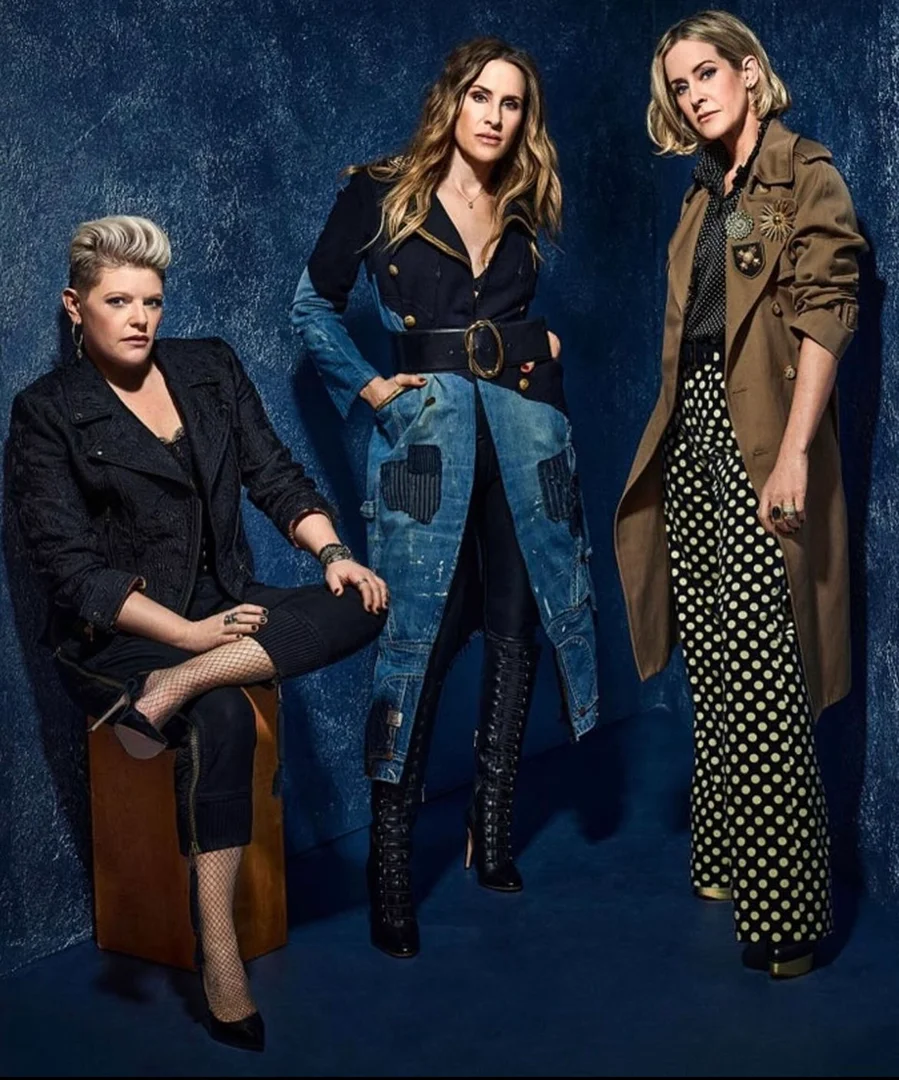 Three women pose in a studio setting against a blue textured wall. One woman sits on a box with fishnet stockings and heels.