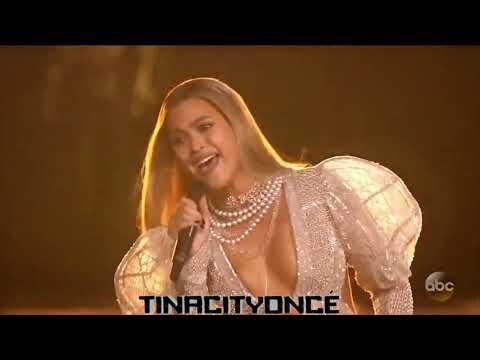 A singing woman with blonde hair and a multi-strand pearl necklace is shown from the chest up against a blurred dark background.