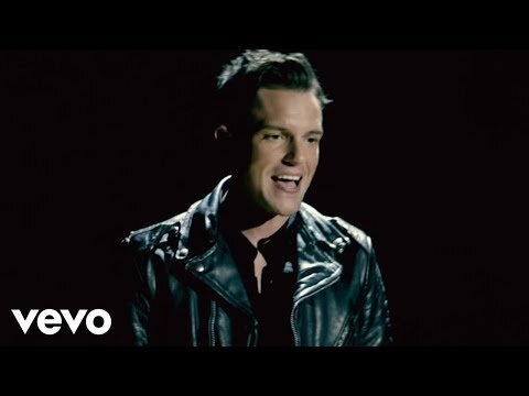 A man singing in a black leather jacket stands against a dark background with dramatic lighting, capturing a moment of musical expression.