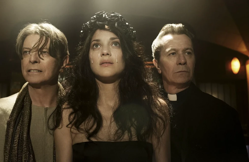 David Bowie, a woman wearing a floral headpiece with tears on her face, and Gary Oldman stand together in an indoor setting.