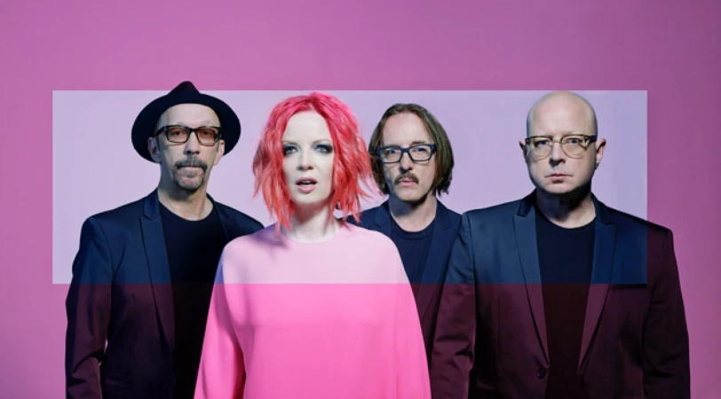 A musical band of four members posing against a pink backdrop; a woman with pink hair stands out in the middle, flanked by three men.