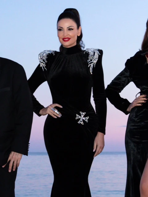Candid shot of a woman in a long black velvet dress with ornate shoulder and waist embellishments, posing on a beach at dusk.
