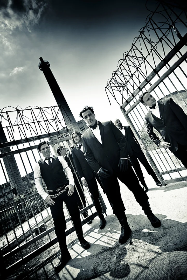 A group of men in dark suits stand in front of a metal gate and industrial buildings, captured with a low angle, black and white shot.