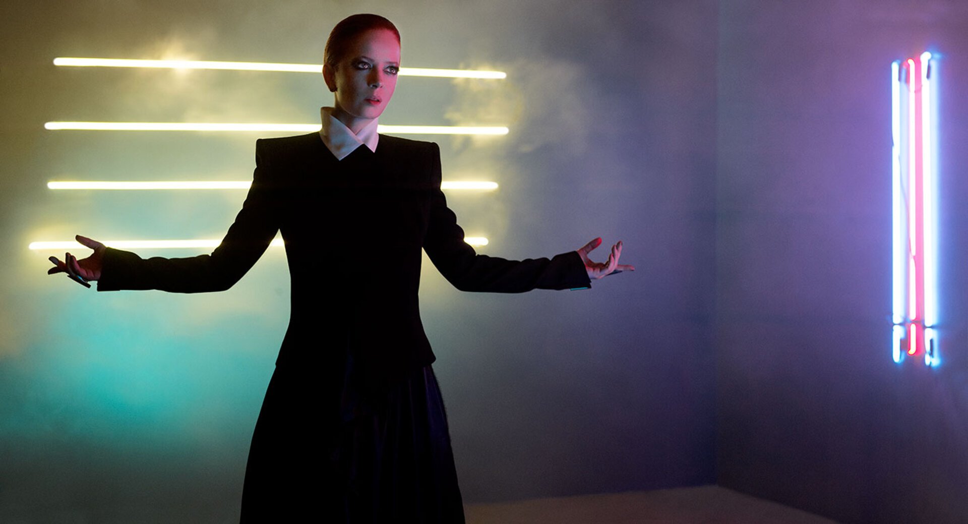 Woman in black dress poses with arms outstretched in a room with neon lights creating a futuristic, fashion-forward and stylized aesthetic.