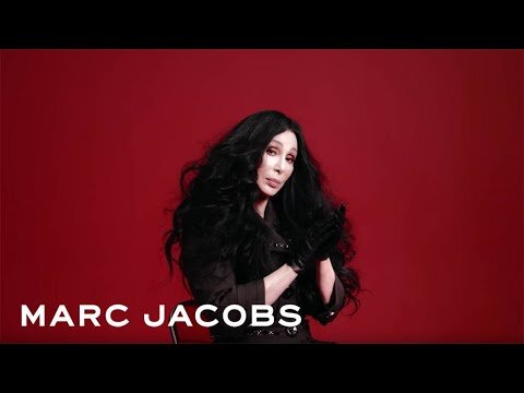 Singer Cher poses in a photo studio against a red backdrop, wearing black gloves and a black jacket over a dark top.