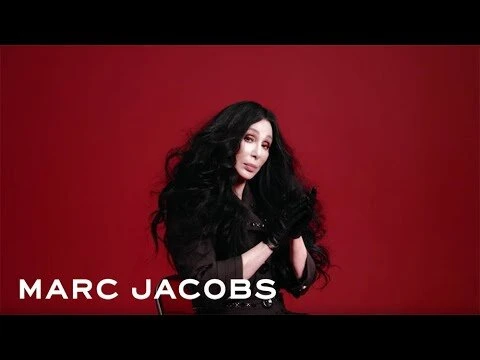 Singer Cher poses in a photo studio against a red backdrop, wearing black gloves and a black jacket over a dark top. 