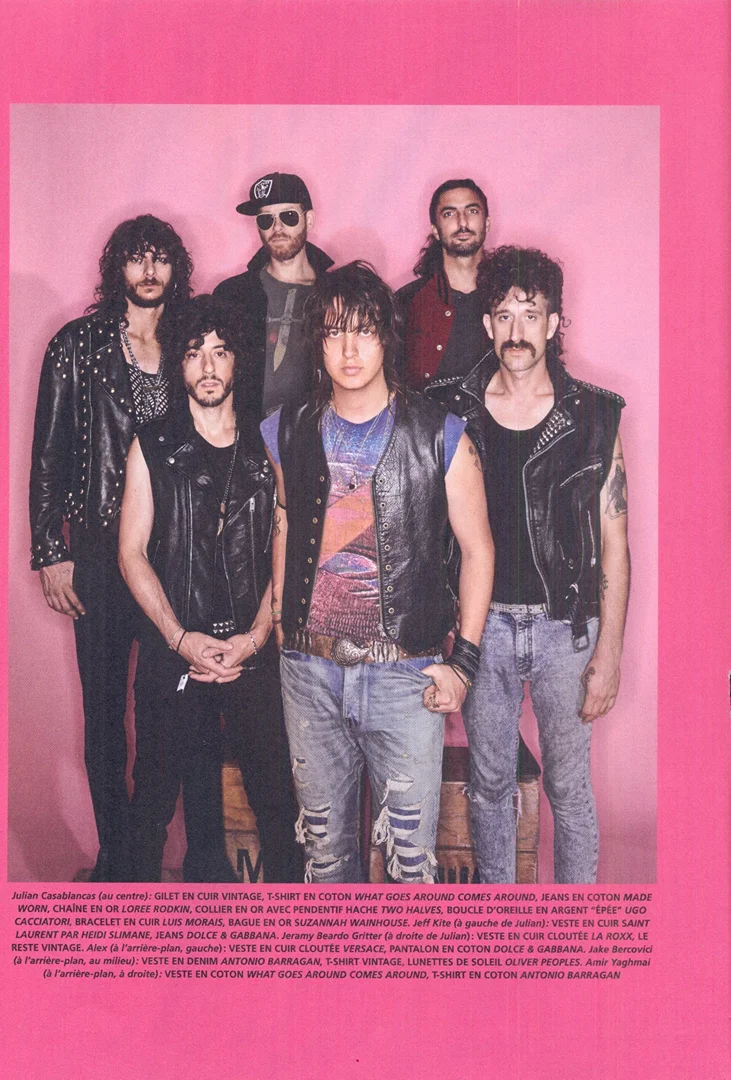 Six adult males posing indoors against a pink backdrop, showcasing leather vests, t-shirts, and jeans, with visible light source.