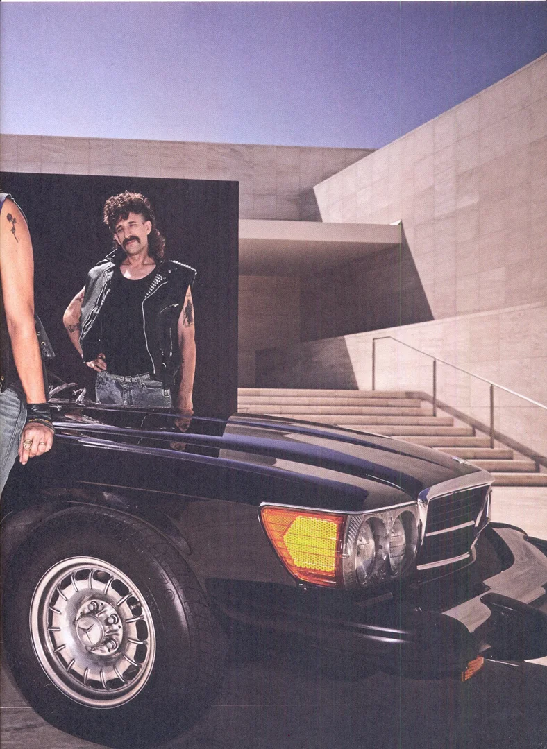 Black vintage car with a man in a black vest and mullet standing in the background in front of a modern architectural building.