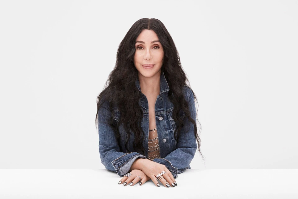 Portrait of Cher in a denim jacket with hands folded on a white table, posing against a white background. Studio lighting, medium shot.