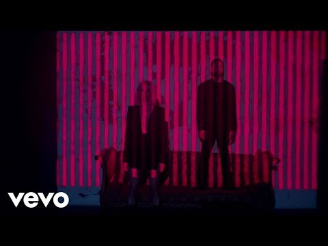 Two adults stand in front of a couch and striped wall in a music video with pink and blue lighting and soft focus.