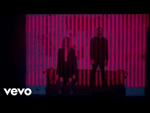 Two adults stand in front of a couch and striped wall in a music video with pink and blue lighting and soft focus.