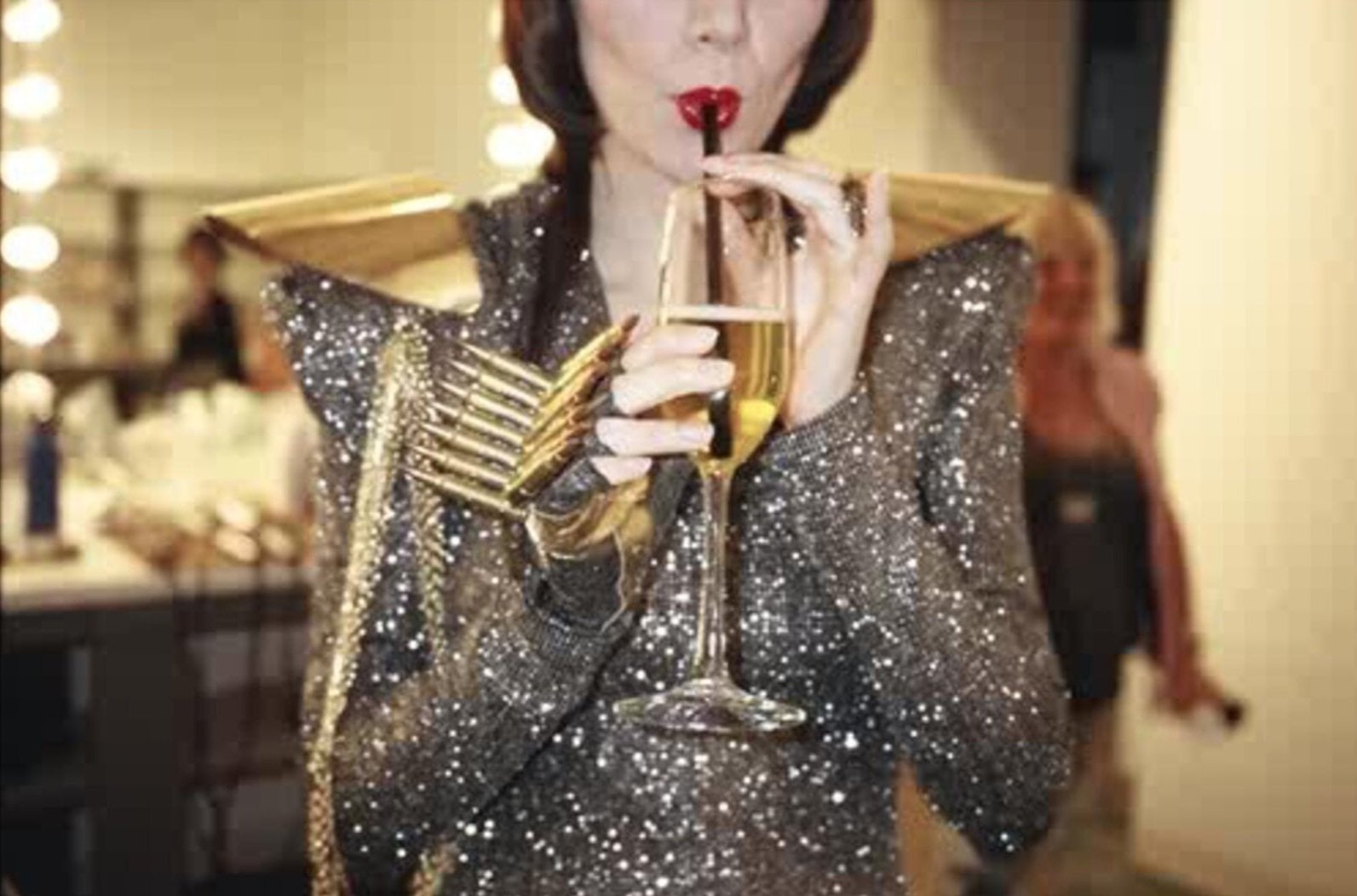 Adult female in a sequin dress drinks champagne through a straw, posing in what appears to be a photo studio setting.