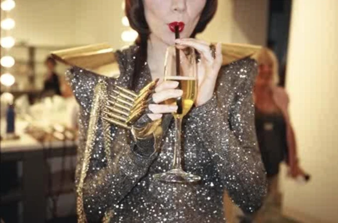 Adult female in a sequin dress drinks champagne through a straw, posing in what appears to be a photo studio setting.
