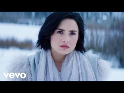 Close-up of a woman with short dark hair and light skin wearing a gray sweater and scarf in a snowy outdoor environment.