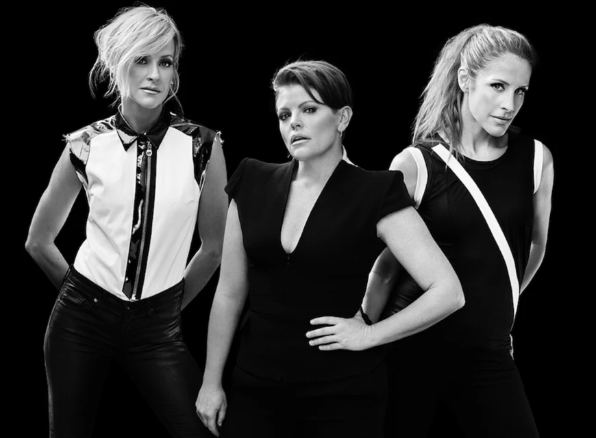 Black and white studio shot of three adult women with dark hair and light skin wearing fashionable black and white dresses against black background.
