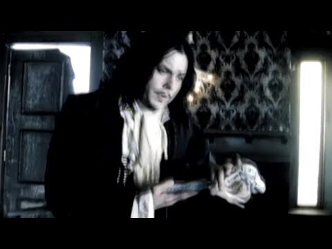 A man with long dark hair, wearing a dark jacket and ruffled white shirt, holds a vintage microphone in a dimly lit room.