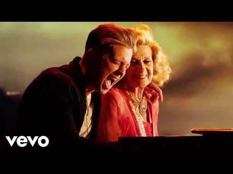 A male vocalist and a senior woman wearing a red dress are smiling at each other. The scene is brightly lit and looks stage-like.