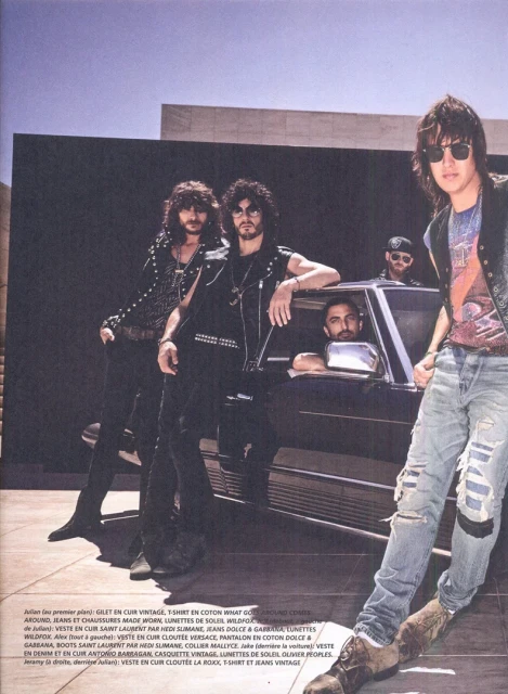 A group of five stylish men pose in front of a black car and modern building. The photo has a vintage editorial fashion style.