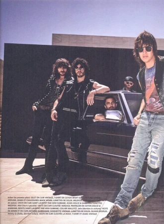 A group of five stylish men pose in front of a black car and modern building. The photo has a vintage editorial fashion style.