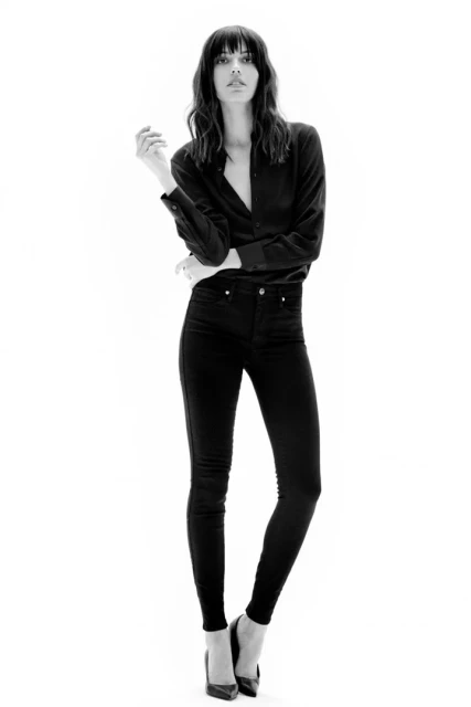 Full-length studio shot of a brunette woman with bangs in a black button-down shirt, black jeans, and black heels on a white background.