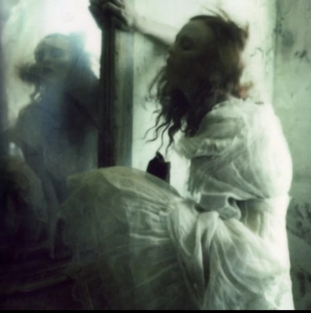 Artistic, blurred image of a woman in a white dress gazing into a vintage mirror with visible wall texture in a shadowy indoor space.