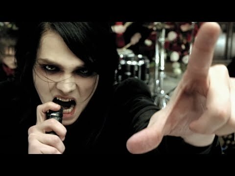 Close-up of a man with dark hair singing into a microphone and reaching towards the camera with styled clothing.