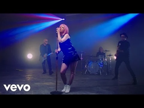 A singer with pink hair performs on stage with a band, bathed in blue and purple stage lights, for a music video. The singer stands at a microphone.
