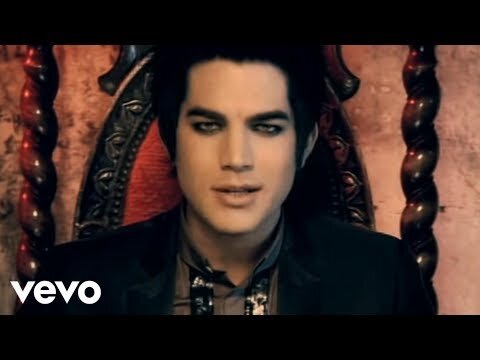 Close-up portrait of an adult male with dramatic eye makeup, wearing a black suit, against a dark, ornate background with vevo logo.