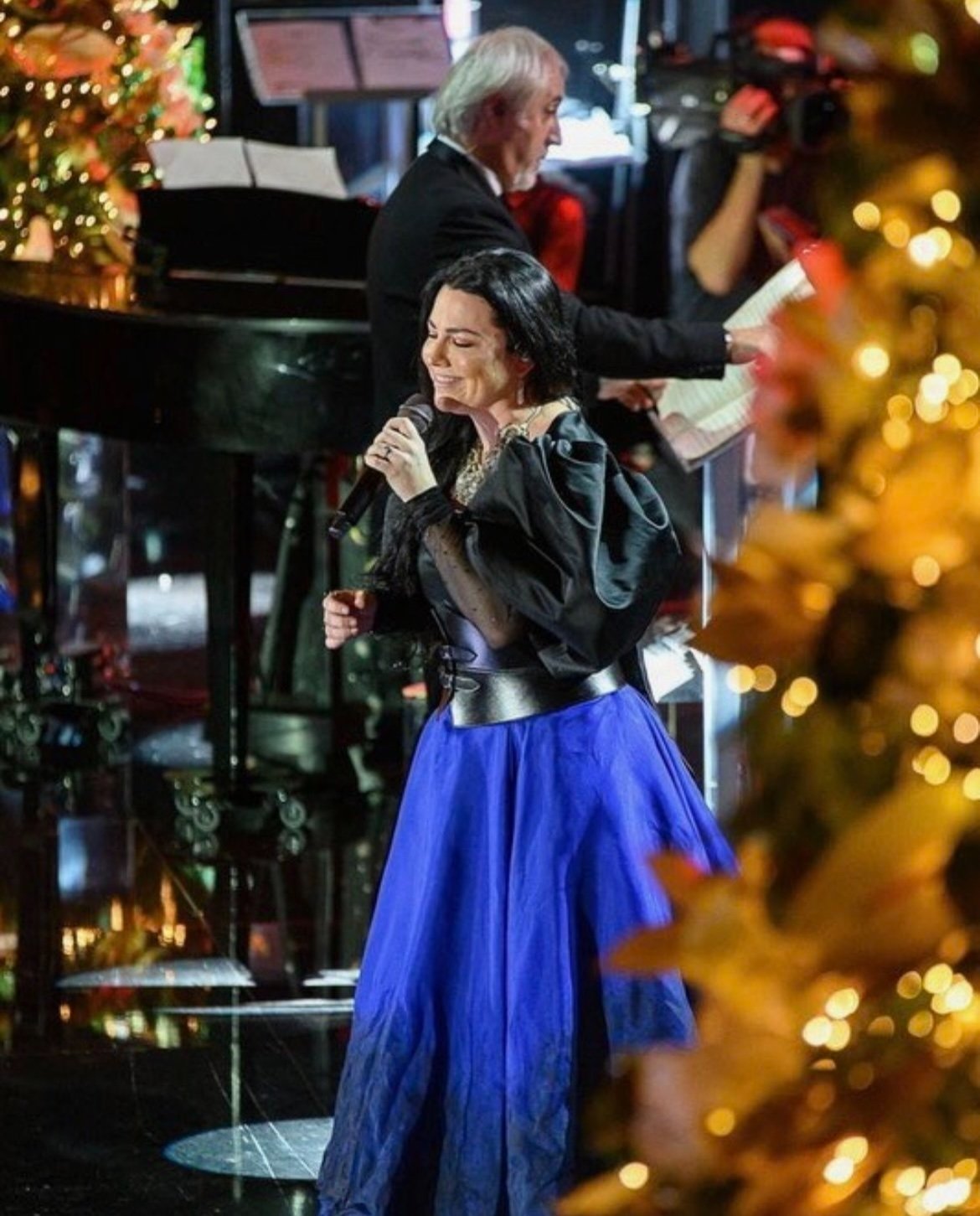 A woman in a black top and blue skirt sings into a microphone with a pianist and Christmas tree in the background on stage.