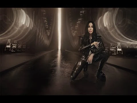 Cher in a futuristic, dark, and stylish setting. The pop singer is captured in a crouching pose, wearing a sleek, black outfit with platform shoes.