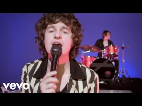 Singer performing into a microphone with a drummer visible in the background against a blue backdrop, bright studio lighting.