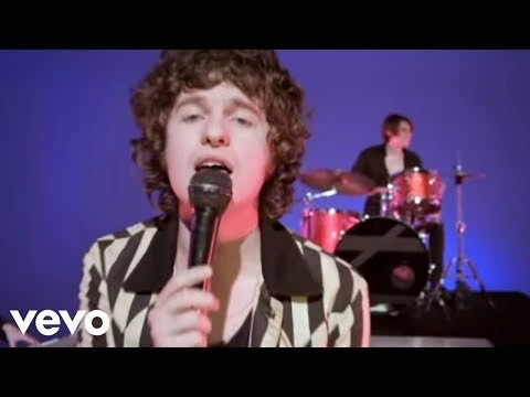 Singer performing into a microphone with a drummer visible in the background against a blue backdrop, bright studio lighting.