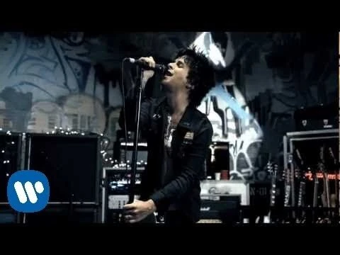 A singer performs on stage with a microphone in a dark music venue with amplifiers and graffiti art in the background.