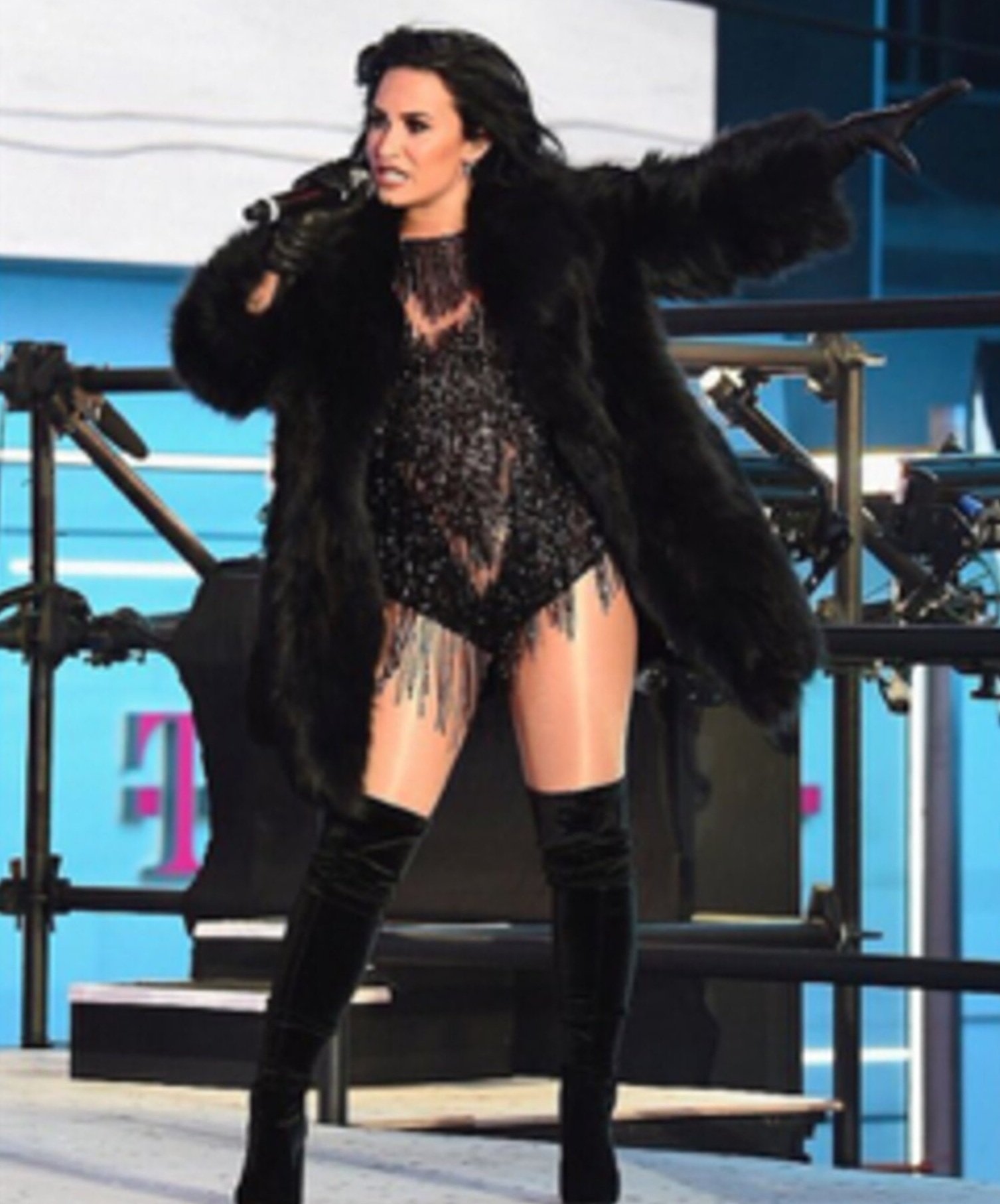 A solo singer with dark hair performs on stage, wearing a black fur coat over a sparkly bodysuit and thigh-high boots, holding a microphone.