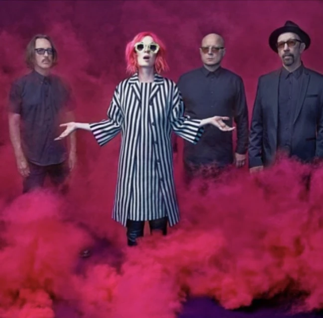 A band of four members stands in front of a pink smoke background. A woman with pink hair is in the center wearing sunglasses.