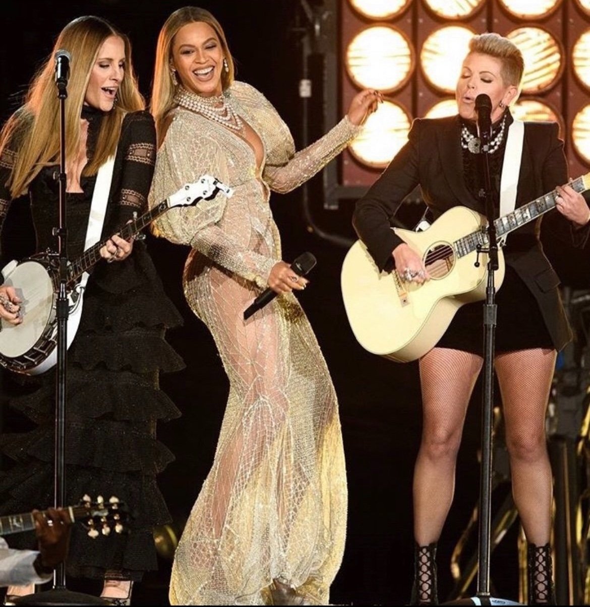 Three female musicians perform on stage: Beyoncé in a shimmering gown singing with a microphone, and two women playing stringed instruments.