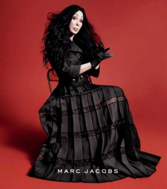 Portrait of Cher sitting in a black dress and gloves against a red backdrop, with the Marc Jacobs logo visible at the bottom.