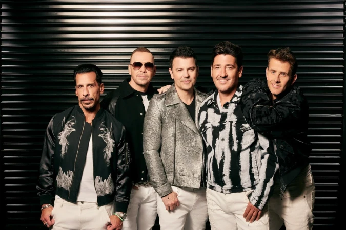 Five men posing together against a dark corrugated metal shutter background, styled by Marjan Malakpour.