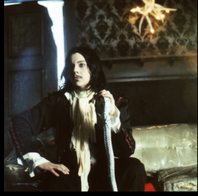 Person in dark clothing poses indoors with a snake draped over their arm, looking off-camera. Wallpapered room with chandelier.