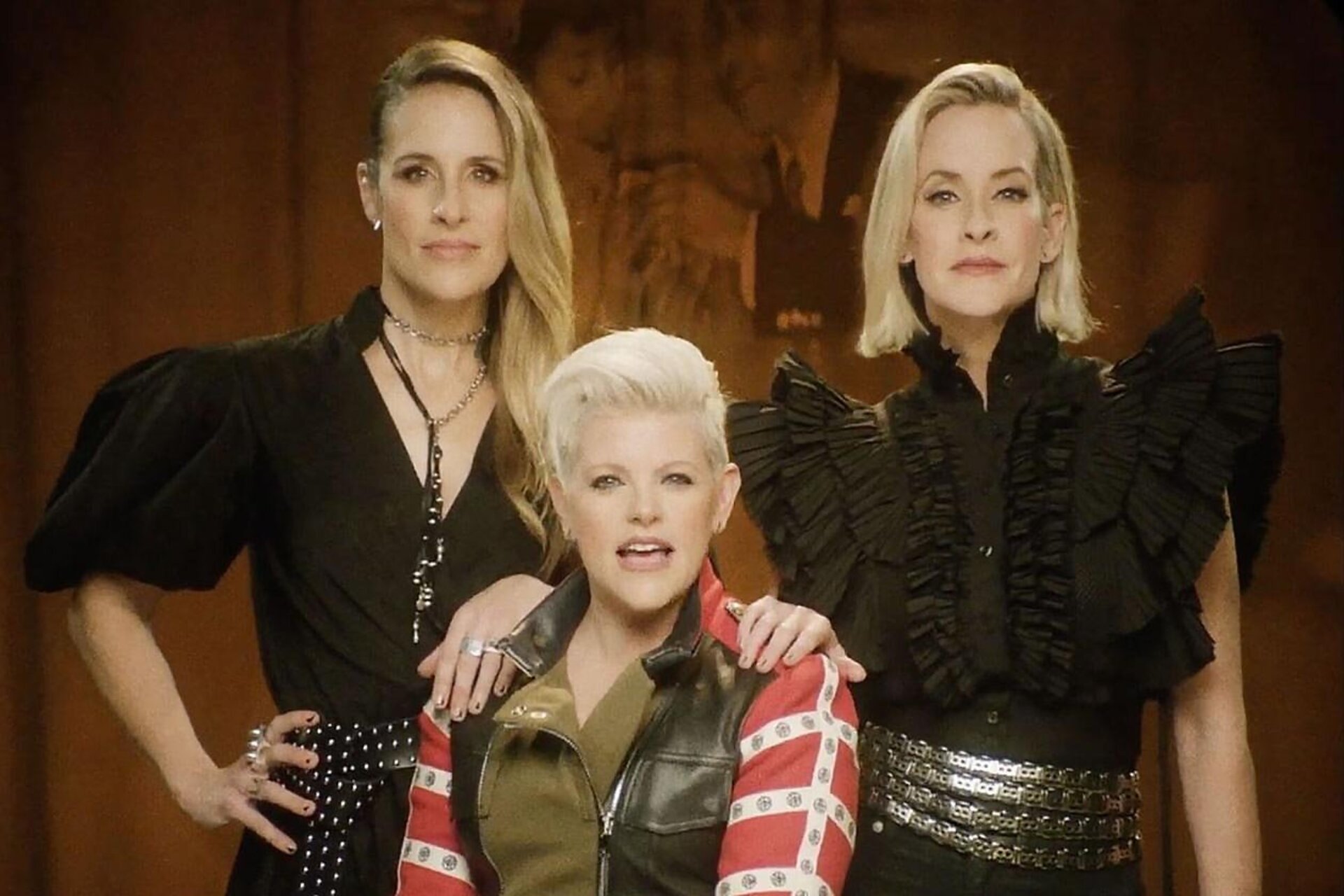 Three women pose indoors against a neutral backdrop. The woman in the center wears a colorful jacket, while the women on either side wear black tops.