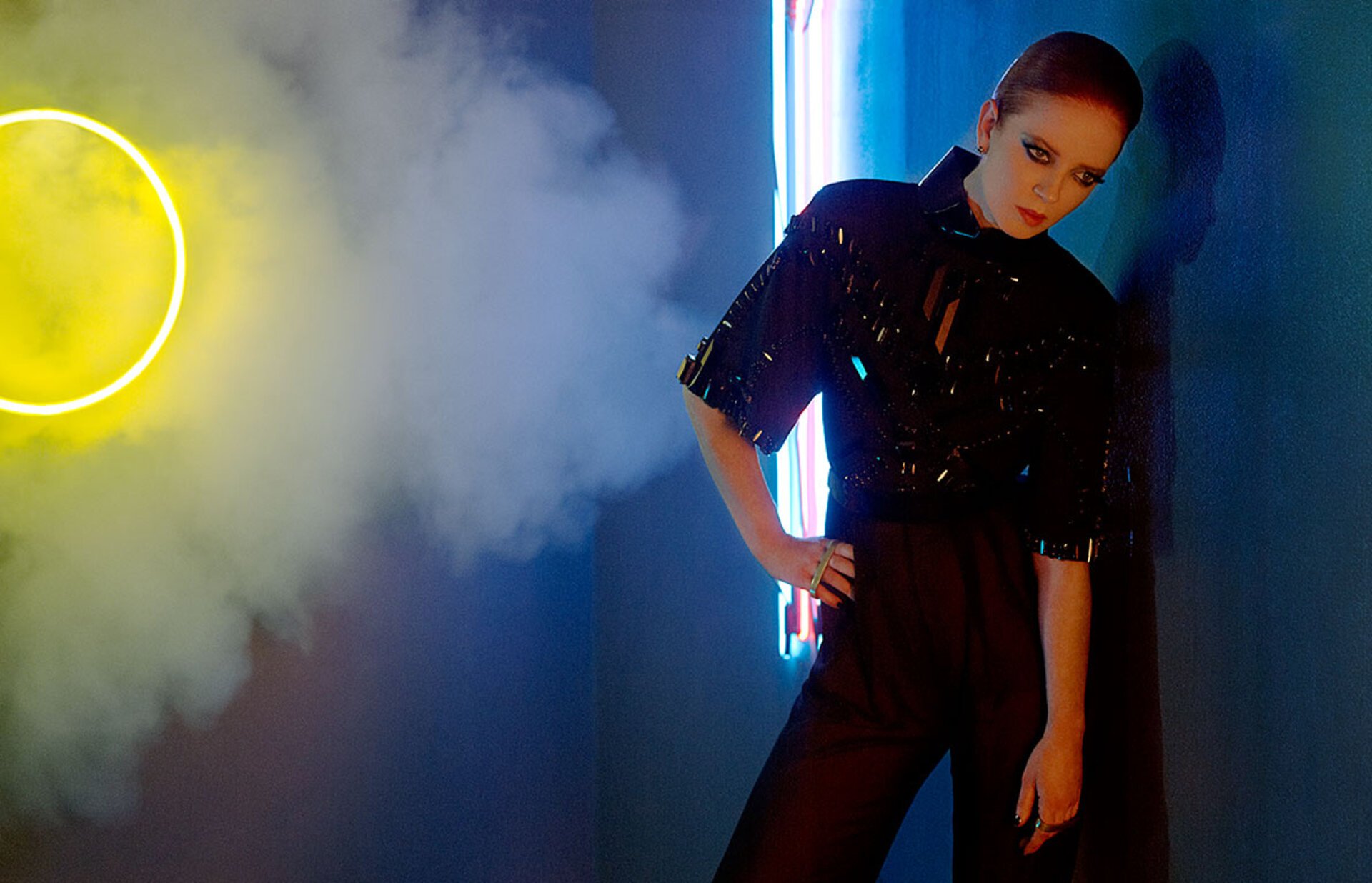 Stylish woman in black top and trousers poses in a dimly lit studio with neon lights and smoke, creating a fashionable and atmospheric scene.