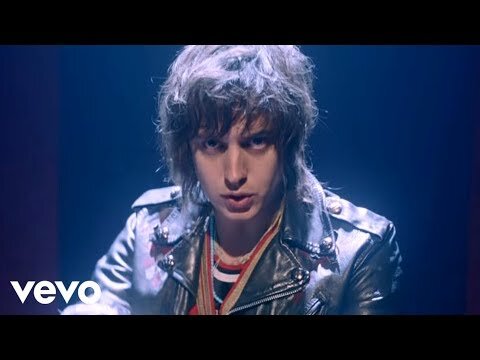 Close-up of a fair-skinned person with messy brown hair in a silver jacket, looking at the camera in a music video scene.