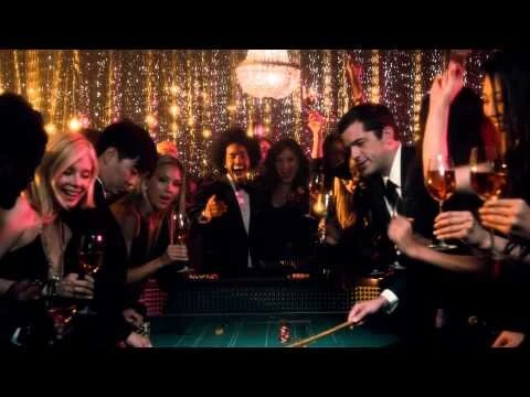 A group of elegantly dressed people laugh and cheer around a casino table with a roulette dealer and champagne at a festive party.