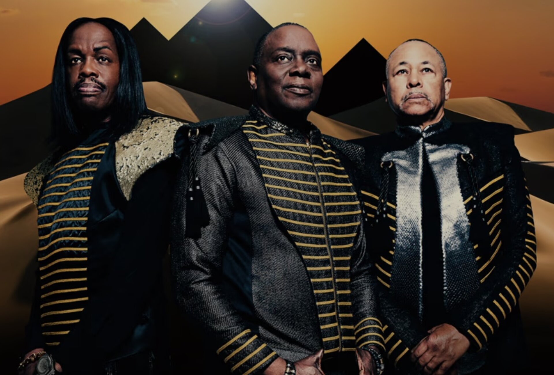 Three adult male musicians in black and gold striped jackets stand in front of a desert landscape with pyramids in the background.